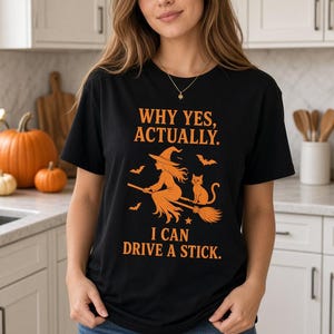 Può includere: Una t-shirt nera a maniche corte con testo grafico arancione che recita "WHY YES, ACTUALLY. I CAN DRIVE A STICK." Il design include un'illustrazione di una strega a cavallo di una scopa con un gatto e diversi pipistrelli attorno alla figura.