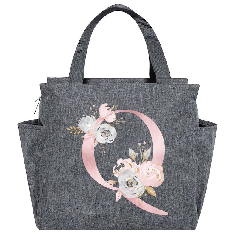 May include: Gray tote bag with a floral design and the letter "Q". The bag has a handle, side pockets, and a zipper closure. The floral design includes pink, white, and gold flowers.