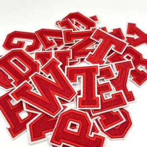Op de afbeelding: Een verzameling rode vilt letters met witte randen. De letters zijn in een vetgedrukte, collegiale stijl, verspreid over een witte ondergrond. Ideaal voor het personaliseren van kleding of knutselwerkjes, deze patches bieden een klassieke, gepersonaliseerde touch.