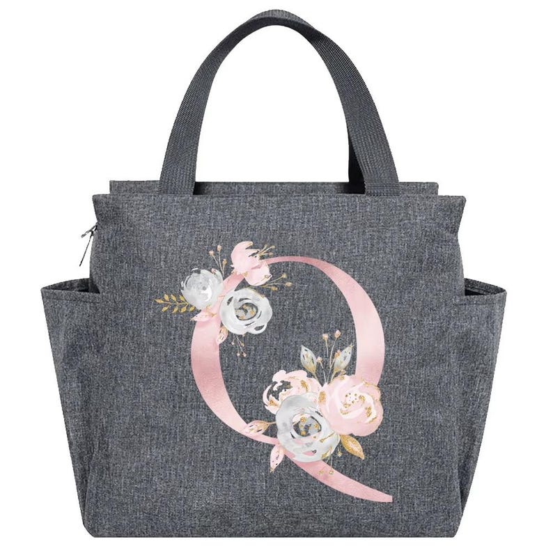 May include: Gray tote bag with a floral design and the letter "Q". The bag has a handle, side pockets, and a zipper closure. The floral design includes pink, white, and gold flowers.