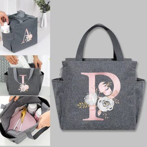 Pode incluir: Saco de almoço cinzento isolado com uma grande letra "P" rosa e design floral. O saco tem uma pega superior, bolsos laterais e um fecho de correr. Concebido para transportar alimentos e bebidas.