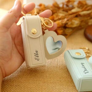 May include: A light beige leatherette keychain case with the name "Ella" printed on it. The case has a gold-colored button closure and is attached to a gold-colored keychain with a heart-shaped charm. A second, similar case in light blue with the name "Daisy" is also visible.