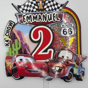 May include: A colorful birthday cake topper featuring Lightning McQueen and Mater from the Cars movie. The topper includes the name "Emmanuel", the number "2", Route 66 signage, and checkered flags. The background has a desert landscape.
