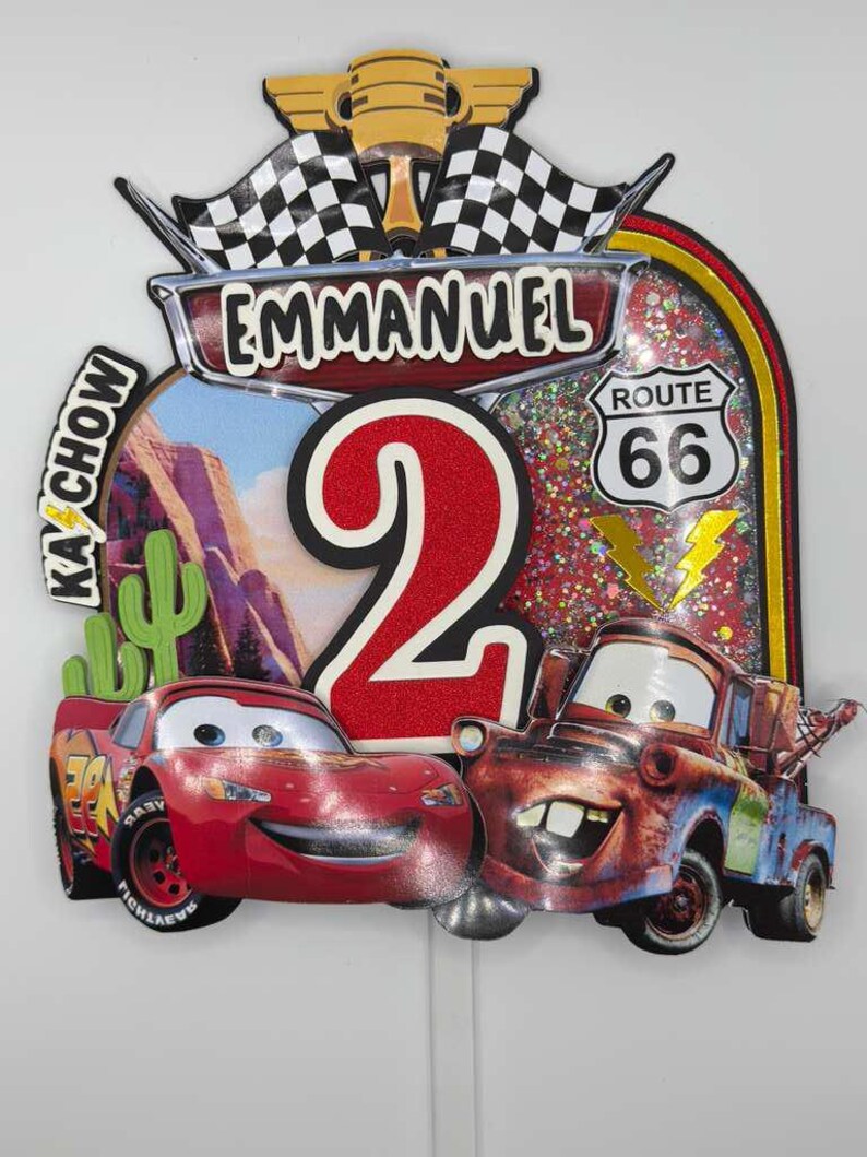 May include: A colorful birthday cake topper featuring Lightning McQueen and Mater from the Cars movie. The topper includes the name "Emmanuel", the number "2", Route 66 signage, and checkered flags. The background has a desert landscape.