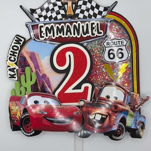 May include: A colorful birthday cake topper featuring the name "Emmanuel" and the number "2". The design includes images of Lightning McQueen and Mater from the movie "Cars", along with Route 66 signage and checkered flags.
