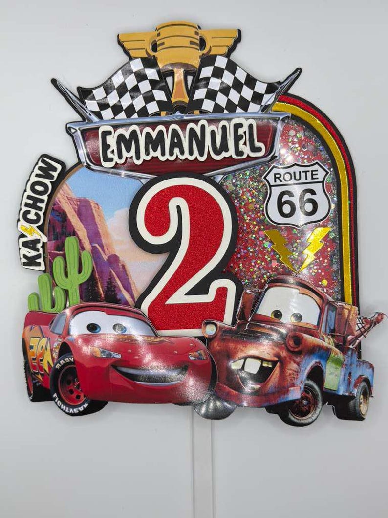 May include: A colorful birthday cake topper featuring the name "Emmanuel" and the number "2". The design includes images of Lightning McQueen and Mater from the movie "Cars", along with Route 66 signage and checkered flags.