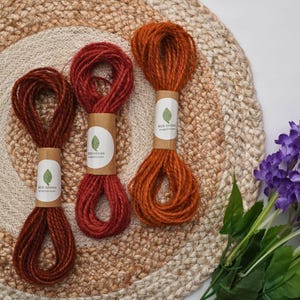 May include: Three bundles of twisted twine in shades of dark red, medium red, and orange. Each bundle is secured with a tan paper band featuring a white circular label with a green leaf icon and the text ECO GROWN ecogrown.com printed on it.