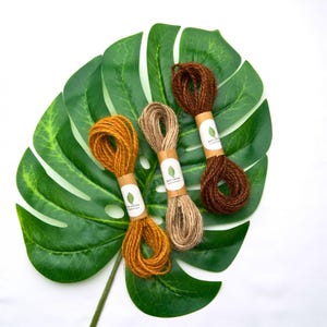 May include: Three bundles of twisted jute twine in shades of mustard yellow, natural tan, and dark brown. Each bundle is secured with a white paper band featuring a green leaf logo and the text Eco Grown, arranged on a large green leaf.