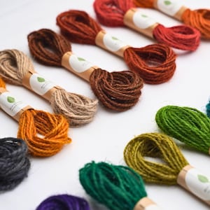 May include: A collection of coiled jute twine bundles in various colors including red, orange, brown, tan, yellow, green, and purple. Each bundle is secured with a tan paper band featuring a small green leaf icon and the text ECO GREEN.