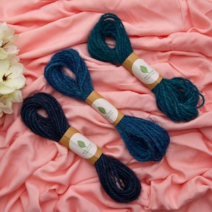 May include: Three bundles of dark teal jute twine, each secured with a tan paper label featuring a green leaf icon and the text ECO GROWN. The twine is textured and fibrous, arranged diagonally across a soft, draped peach-colored fabric.