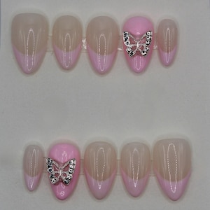 May include: A set of oval-shaped false nails. The nails feature a light pink French tip design with a nude base. Two nails have a silver butterfly embellishment with black and clear rhinestones.