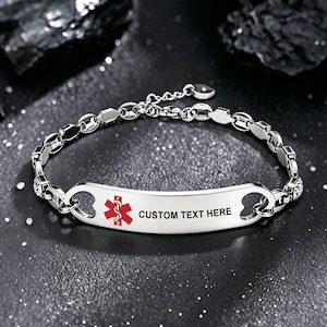 Könnte beinhalten: Ein silberfarbenes medizinisches Notfallarmband mit einer polierten Metallplatte, einem roten Star of Life-Symbol und dem schwarzen Text CUSTOM TEXT HERE. Das Armband besteht aus einer Kette mit ovalen Gliedern und einem kleinen herzförmigen Anhänger.
