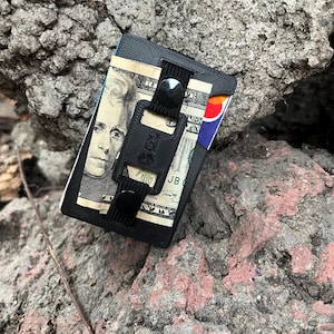 May include: A black minimalist wallet featuring a textured surface, a central elastic strap with a black snap button, and a bottle opener attachment. The wallet holds a twenty-dollar bill and a credit card with visible red, yellow, and blue colors.