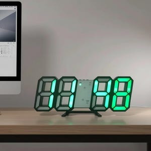 Puede incluir: Un reloj digital con marco negro y números LED verde brillante. El reloj muestra la hora en un estilo moderno y minimalista. Está sobre una superficie de madera, con un monitor de ordenador visible en el fondo.