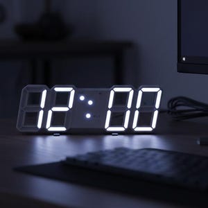 May include: A digital clock displaying "12:00" with bright white LED lights. The clock is on a desk with a computer monitor, keyboard, and mouse. The background is dark, creating a contrast with the illuminated clock.