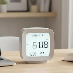 Puede incluir: Un despertador digital cuadrado gris muestra la hora 6:08, la fecha 2022.10.30, la humedad al 69% y la temperatura a 53 grados. El reloj está sobre un escritorio de madera.
