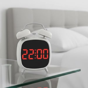 May include: A white digital alarm clock displaying 22:00 in red numerals. The clock sits on a glass table, with a bed and a small plant in the background. The clock has a retro design with two bells on top.