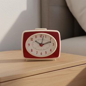 May include: A rectangular analogue alarm clock with a cream-coloured frame and a deep red face. The clock features a white oval dial with black numerals from one to twelve, black hour and minute hands, and a small red centre point for the hands.