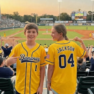 Op de afbeelding: Twee mensen dragen gele honkbalshirts. Eén shirt heeft het woord "Bananas" en het nummer "1" op de voorkant. De andere heeft "Jackson" en "04" op de rug. Ze zijn bij een honkbalwedstrijd.