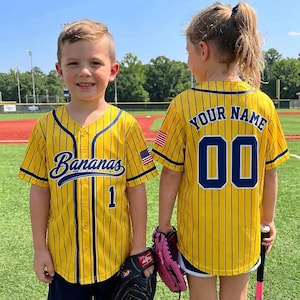 Op de afbeelding: Twee kinderen dragen gele honkbalshirts met marineblauwe krijtstrepen. Het shirt links zegt "Bananas" en heeft nummer "1". Het shirt rechts zegt "YOUR NAME" en heeft nummer "00". Beide shirts hebben een Amerikaanse vlag patch.