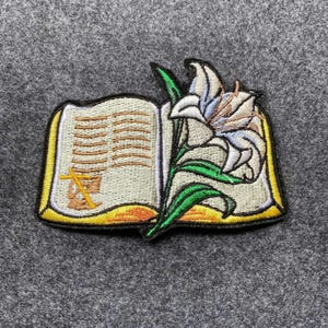 Puede incluir: Parche bordado con un libro abierto con páginas doradas y borde amarillo. Una cruz y un cáliz dorados son visibles en la página izquierda. Un lirio blanco con hojas verdes está a la derecha.