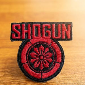 May include: Embroidered patch with the word "SHOGUN" in red lettering above a circular emblem. The emblem features a red floral design within a black border, set against a wooden surface.