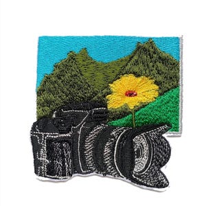 May include: Embroidered patch featuring a black camera with a lens, set against a backdrop of green mountains, a yellow flower, and a blue sky. The patch is detailed with various textures and colors, suitable for photography enthusiasts.