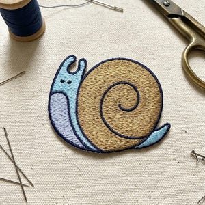 May include: Embroidered snail patch with a light blue body and a tan shell. The patch is surrounded by sewing tools, including needles, scissors, and a spool of blue thread. The snail's shell features a detailed spiral design.