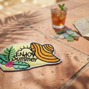 Op de afbeelding: Geborduurde patch met de woorden "Enjoy Summer" en een ontwerp van zon, hoed en bladeren. Een glas ijsthee met munt, een paspoort en zeeglas zijn ook zichtbaar op een bakstenen oppervlak.