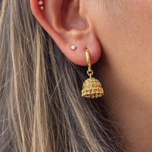 以下が含まれることがあります： 金色のベル型のデザインのイヤリングのクローズアップ。イヤリングは小さなフープから吊り下げられています。耳には2つの小さな輝くスタッドがあります。