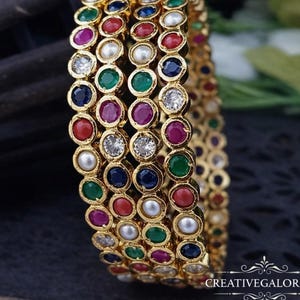 Op de afbeelding: Gouden armbanden met rijen kleurrijke edelstenen, waaronder rood, blauw, groen en paars, afgewisseld met parels en heldere kristallen. De armbanden zijn gestapeld en tonen het ingewikkelde ontwerp en de levendige kleuren.