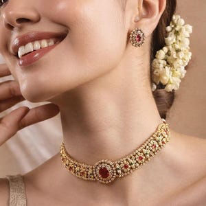 以下が含まれることがあります： 赤と緑の宝石が付いた、ゴールドトーンのネックレスとイヤリングのセット。 ネックレスは、中央に大きな石が付いたチョーカータイプです。 イヤリングは、中央に赤い石が付いた丸いスタッドです。 画像には「CREATIVEGALOREIN」というテキストが含まれています。