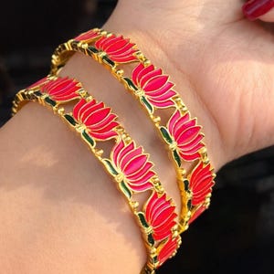 May include: Gold-toned bangles with a repeating lotus flower design in red and pink enamel. The floral details are outlined in black and green. The bangles are worn on a wrist.