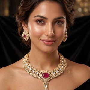 May include: A gold-toned necklace and matching stud earrings set featuring intricate metalwork, clear stones, and prominent oval-shaped deep pink cabochon gems. The necklace has a central pendant drop and a repeating pattern of stones.
