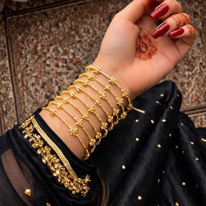 Könnte beinhalten: Mehrere goldene Armbänder schmücken ein Handgelenk, jedes mit zarten floralen Designs. Die Armbänder sind gestapelt, wodurch ein geschichteter Effekt entsteht. Die Fingernägel sind tiefrot lackiert. Der Ärmel eines schwarzen Kleidungsstücks mit goldenen Verzierungen ist sichtbar.