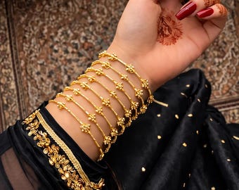 May include: A set of seven thin gold bangles featuring small, evenly spaced spherical accents. The bracelets are worn on the wrist, paired with a black sleeve that has a wide, ornate gold embroidered cuff with floral patterns and gold sequins.