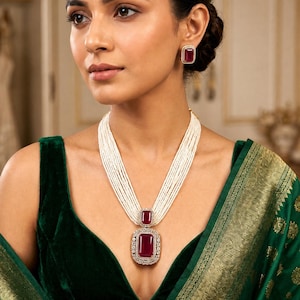 May include: A multi-strand pearl necklace with a large, rectangular ruby pendant and matching earrings. The ruby is surrounded by a halo of small, clear stones. The woman is wearing a dark green velvet top and a green and gold sari.