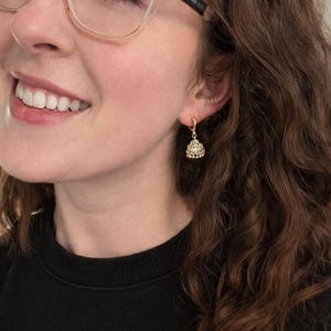 K&ouml;nnte beinhalten: Nahaufnahme eines goldfarbenen Ohrrings mit einem kleinen, dreieckigen Anh&auml;nger. Der Anh&auml;nger ist mit kleinen, funkelnden Steinen besetzt. Der Ohrring ist an einem kleinen Reifen befestigt. Die Person tr&auml;gt ein schwarzes Oberteil.
