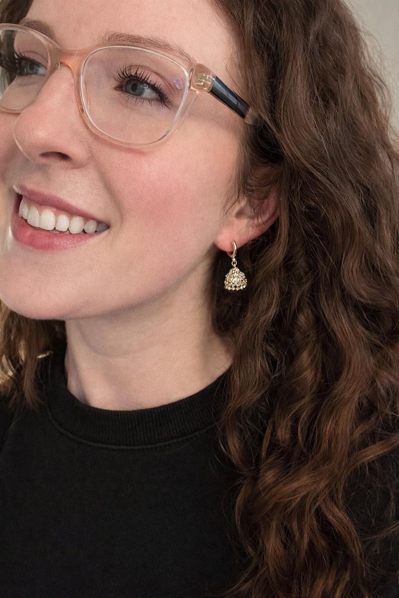 K&ouml;nnte beinhalten: Nahaufnahme eines goldfarbenen Ohrrings mit einem kleinen, dreieckigen Anh&auml;nger. Der Anh&auml;nger ist mit kleinen, funkelnden Steinen besetzt. Der Ohrring ist an einem kleinen Reifen befestigt. Die Person tr&auml;gt ein schwarzes Oberteil.