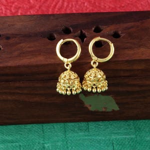 K&ouml;nnte beinhalten: Ein Paar goldfarbene Jhumka-Ohrringe. Jeder Ohrring hat einen Reifen und einen glockenf&ouml;rmigen Anh&auml;nger mit aufwendigen Details und kleinen, herabh&auml;ngenden Perlen. Die Ohrringe werden auf einer dunklen Holzoberfl&auml;che vor einem roten und gr&uuml;nen Hintergrund pr&auml;sentiert.