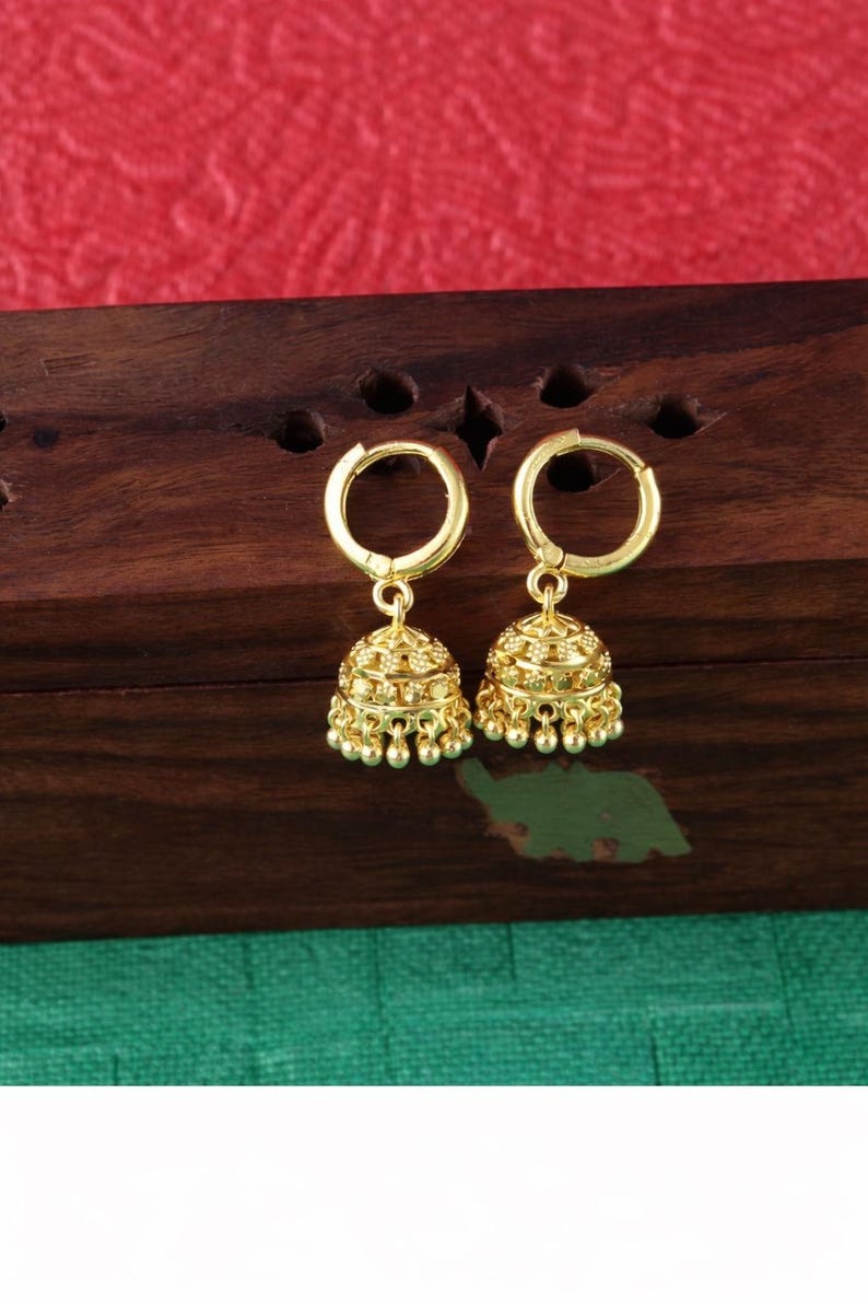 K&ouml;nnte beinhalten: Ein Paar goldfarbene Jhumka-Ohrringe. Jeder Ohrring hat einen Reifen und einen glockenf&ouml;rmigen Anh&auml;nger mit aufwendigen Details und kleinen, herabh&auml;ngenden Perlen. Die Ohrringe werden auf einer dunklen Holzoberfl&auml;che vor einem roten und gr&uuml;nen Hintergrund pr&auml;sentiert.