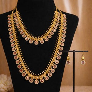 May include: Gold-toned necklace and earring set displayed on a black mannequin. The necklace features multiple strands with circular, red-centered accents. Matching earrings are displayed on a small stand. The jewelry is set against a neutral background.