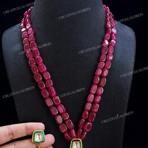 May include: A three-strand ruby necklace and matching earrings. The necklace features large, oval-shaped ruby beads. The earrings have a square green and white top with a gold and pearl drop. The jewellery is displayed on a black mannequin.