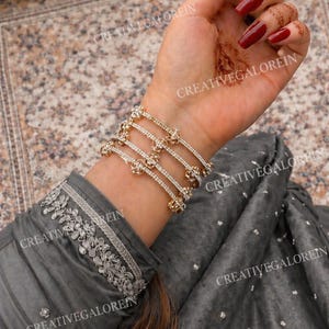 May include: Gold-toned bracelets with floral and rhinestone accents are stacked on a wrist. The bracelets are worn over a gray long-sleeved garment with silver embroidery. The nails are painted a deep red.