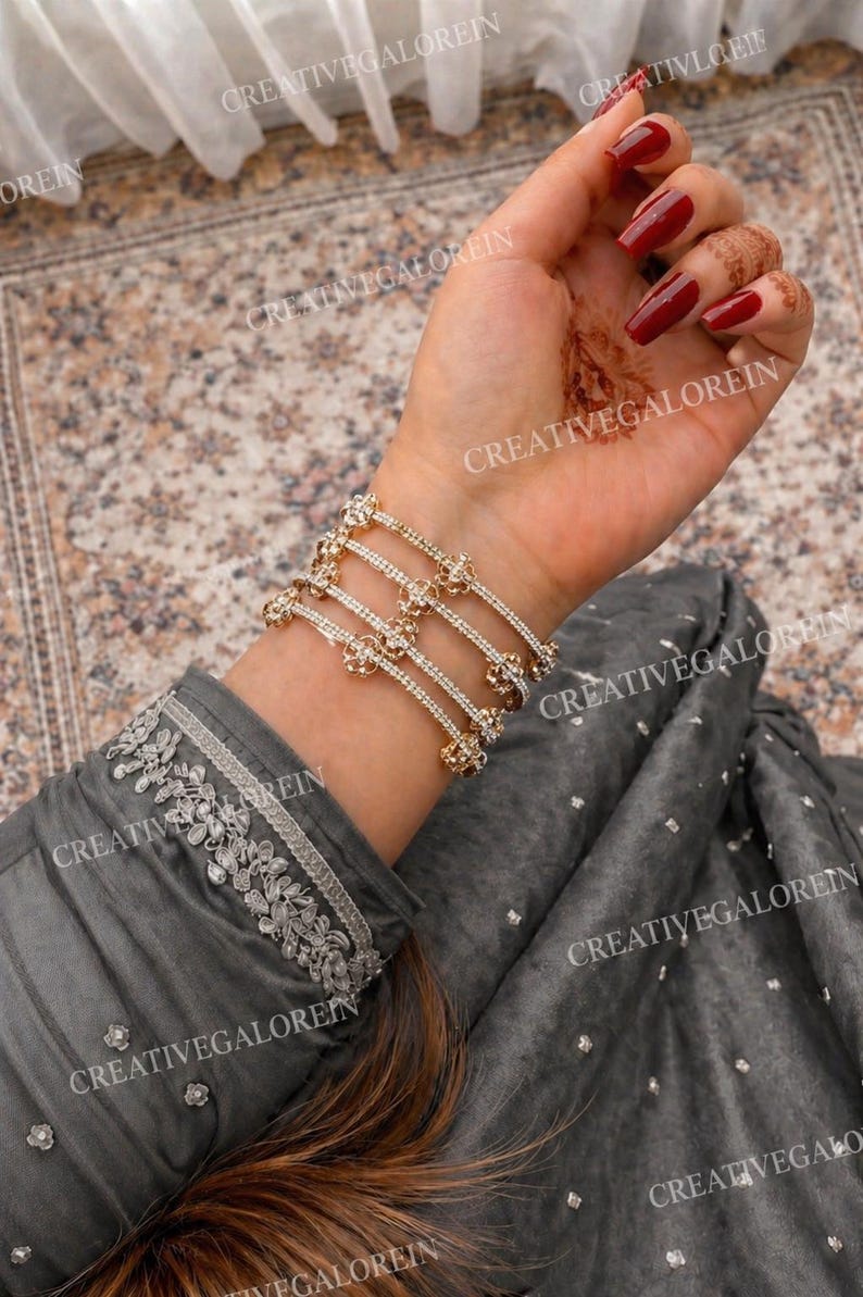 May include: Gold-toned bracelets with floral and rhinestone accents are stacked on a wrist. The bracelets are worn over a gray long-sleeved garment with silver embroidery. The nails are painted a deep red.