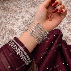 May include: A silver bracelet set with floral designs and clear gemstones. The bracelets are worn on a wrist with red nail polish and henna art. The sleeve of a burgundy garment with silver embroidery is also visible.