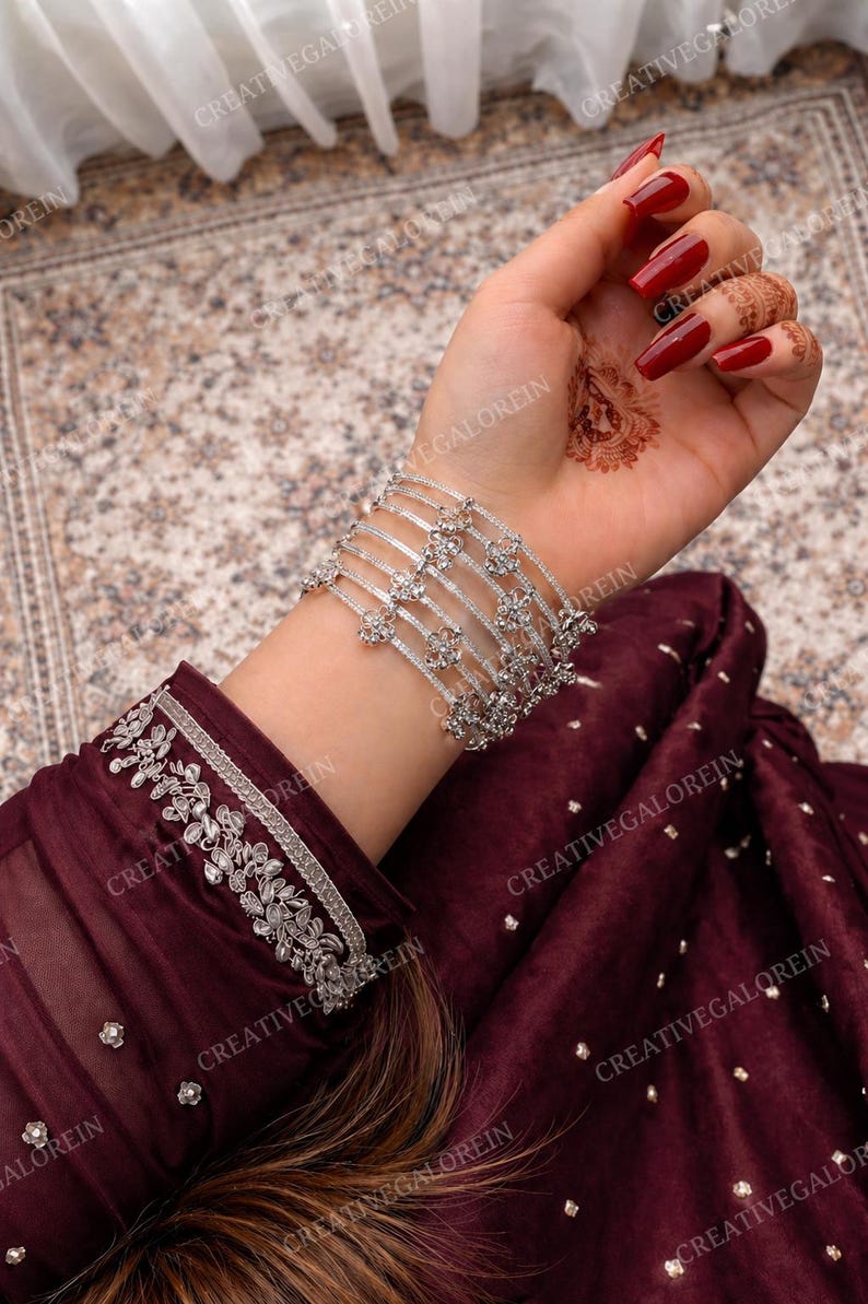 May include: A silver bracelet set with floral designs and clear gemstones. The bracelets are worn on a wrist with red nail polish and henna art. The sleeve of a burgundy garment with silver embroidery is also visible.