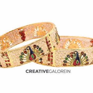 May include: Two gold-colored bangles with intricate designs. Each bangle features a cream-colored band with a peacock motif in blue, green, red, and yellow. The bangles have a filigree pattern and are set against a white background.
