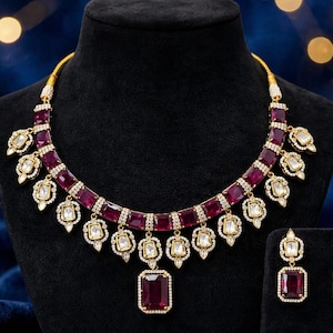 May include: A gold-toned necklace featuring a series of rectangular deep red stones bordered by small white crystals, with dangling teardrop-shaped pendants containing white stones, accompanied by matching rectangular red stone drop earrings.