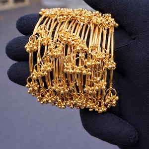 Könnte beinhalten: Eine Sammlung goldener Armreifen, jeder mit kleinen, kugelförmigen Verzierungen. Die Armreifen werden vor einem dunklen Hintergrund präsentiert, der ihr detailliertes Design und ihren strahlenden, goldenen Farbton hervorhebt. Der Schmuck scheint aus massivem Gold zu sein.